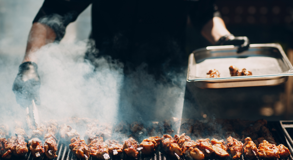 A chef standing at a smoky grill filled with meat