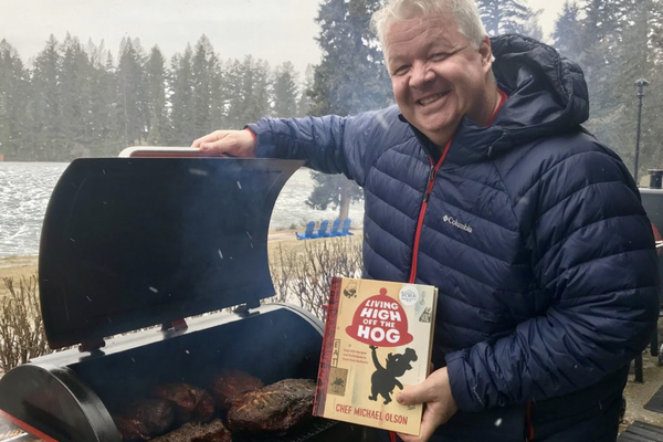 Chef Michael Olson, holding his cookbook in front of a barbecue filled with meat