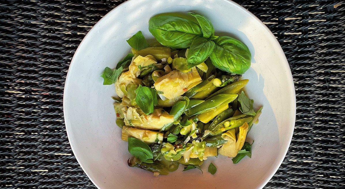 A white bowl with a mix of artichokes, peas and fave beans, garnished with a basil leaf