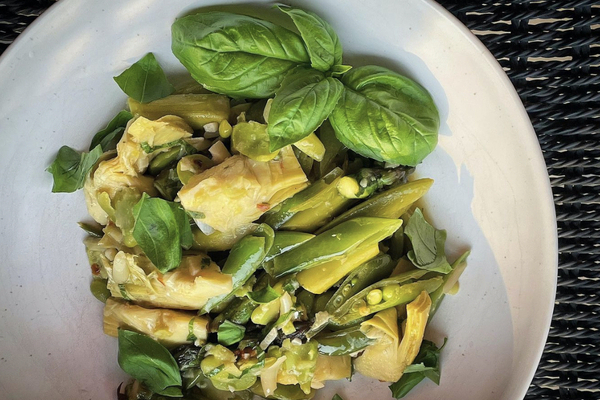 A white bowl with a mix of artichokes, peas and fave beans, garnished with a basil leaf