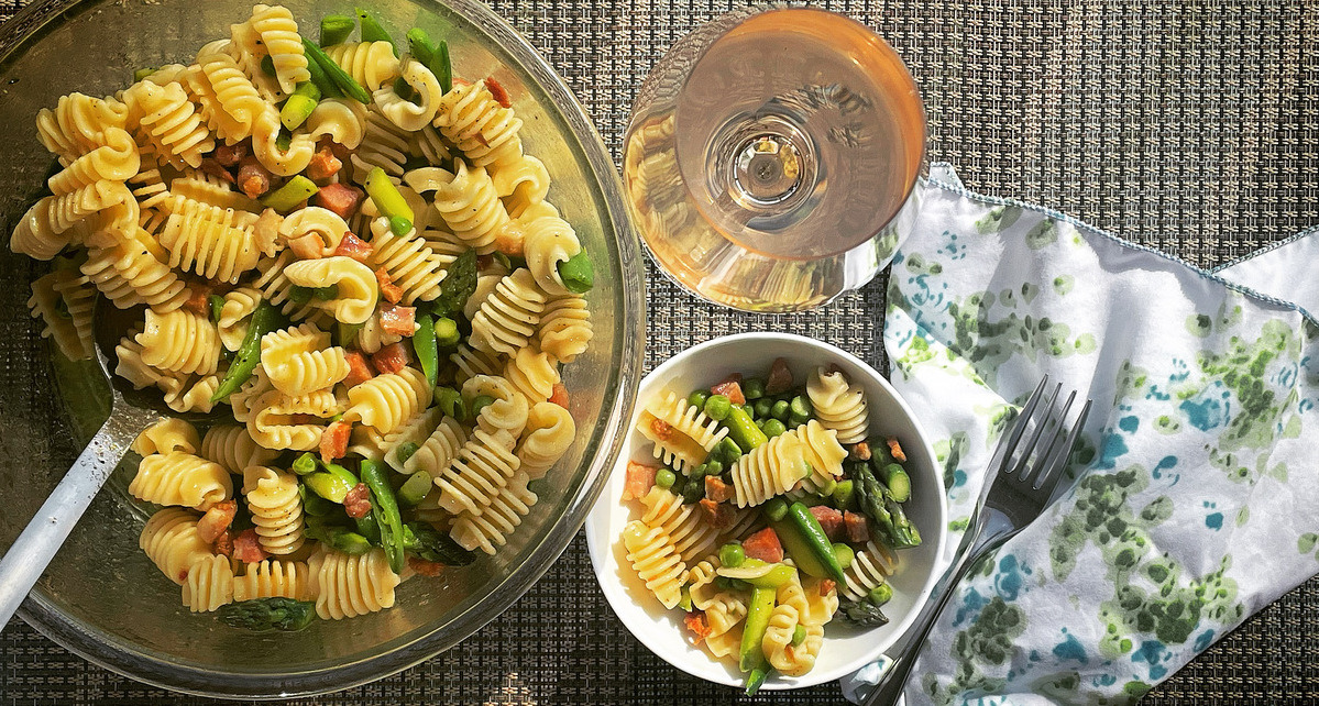 A serving bowl of pasta salad beside a smaller bowl. A floral napkin and glass of rosé are beside it.