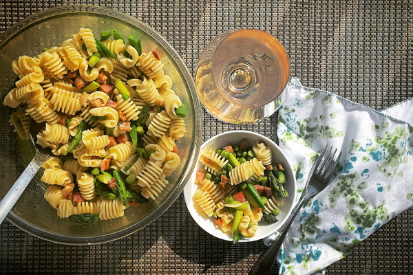 A serving bowl of pasta salad beside a smaller bowl. A floral napkin and glass of rosÃ© are beside it.