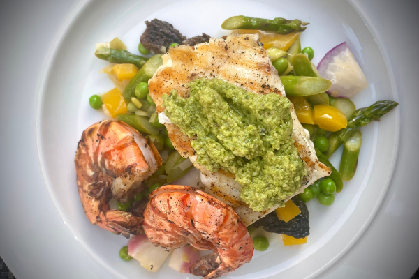 top view of halibut with green garlic scape pesto, colourful vegetables and two pink prawns on a white plate