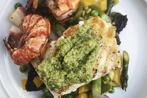 top view of halibut with green garlic scape pesto, colourful vegetables and two pink prawns on a white plate
