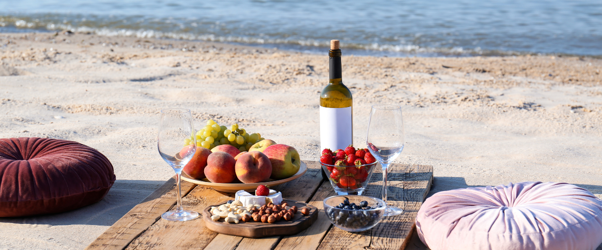 A picnic on a beach with cushions and a bottle of wine and fruit on a wooden plank, the ocean in the background