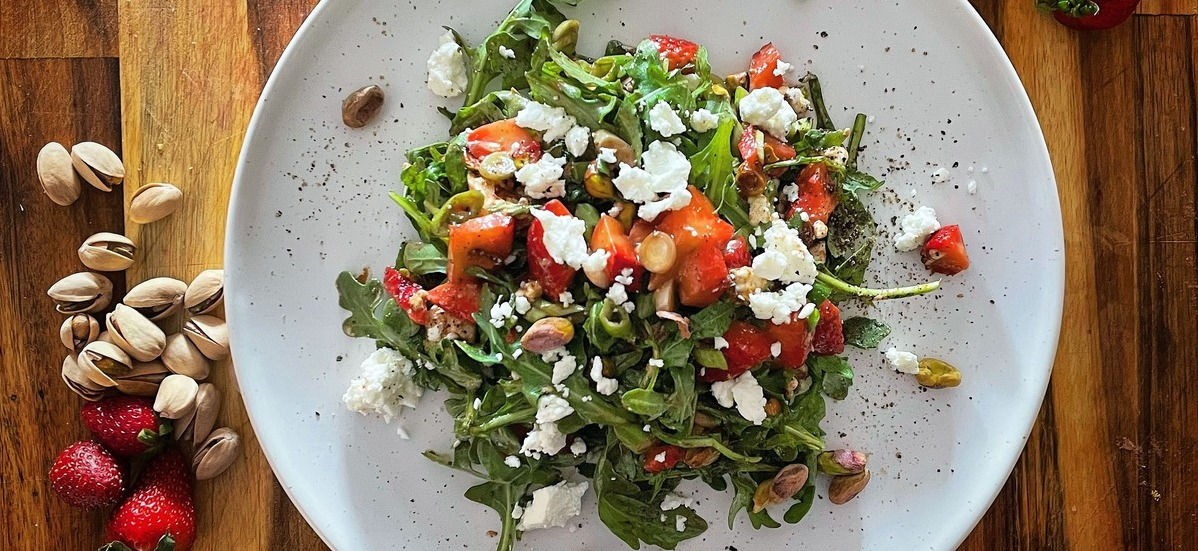 Strawberry and arugula salad on a white plate with strawberries and pistachios scattered around it on a wood board