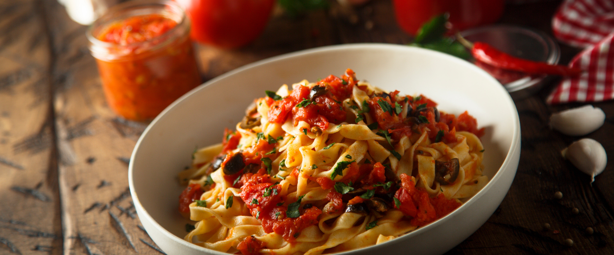 a bowl full of pasta puttanesca on a rustic wood table, herbs and tomatoes scatter around it