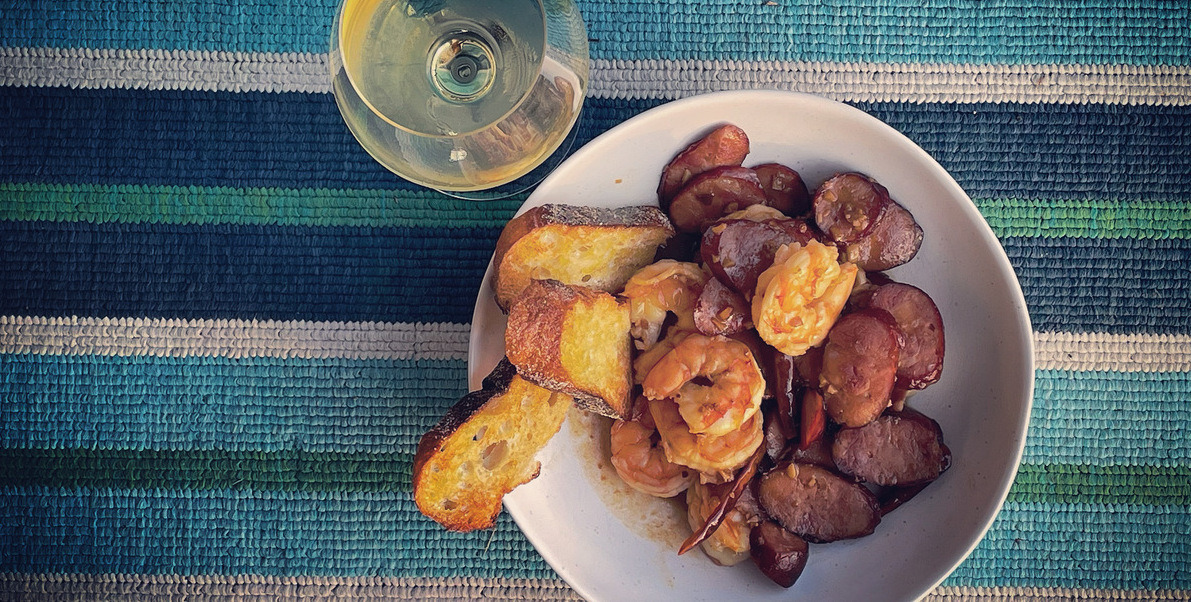 A white bowl filled with shrimp and chorizo with a glass of white wine beside the bowl on a blue striped blanket