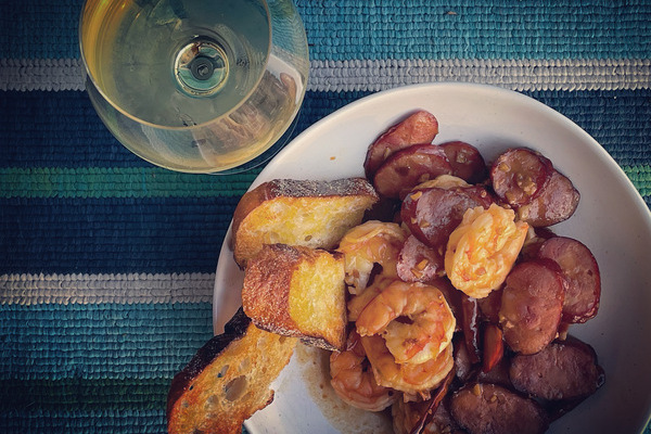 A white bowl filled with shrimp and chorizo with a glass of white wine beside the bowl on a blue striped blanket