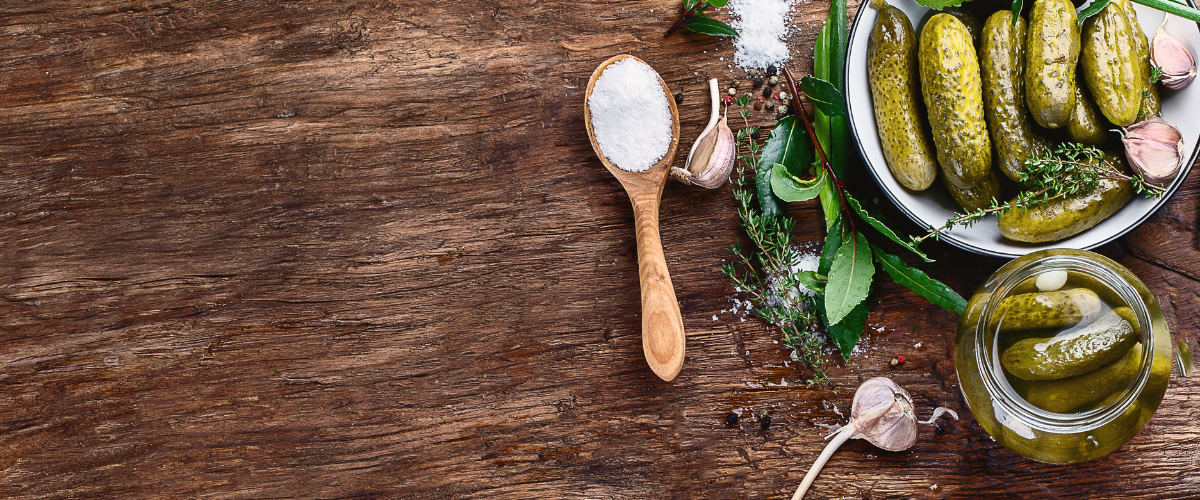 fresh pickles on a wood table surrounded by seasoning, herbs and spices.