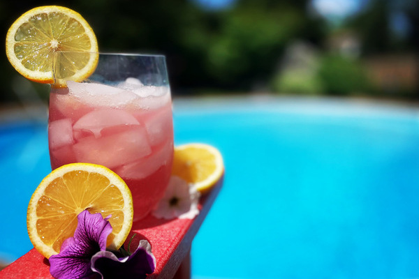 A tumbler full of ice and pink liquid on the arm of a red muskoka chair, surrounded by cut lemon wedges and flowers, in front of a bright blue pool