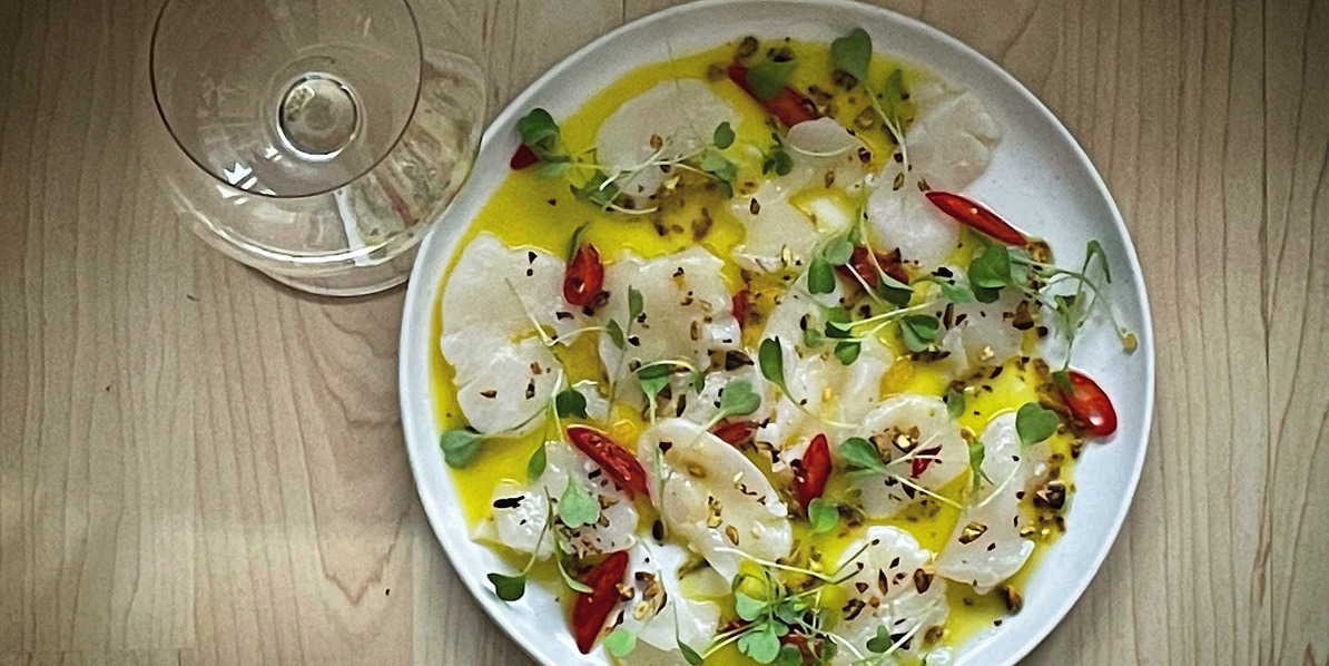 Scallop on a white plate decorated by red chilie peppers, micro greens, a toasted pistachios in a pool of lemon oil. Beside the dish is a glass of white wine