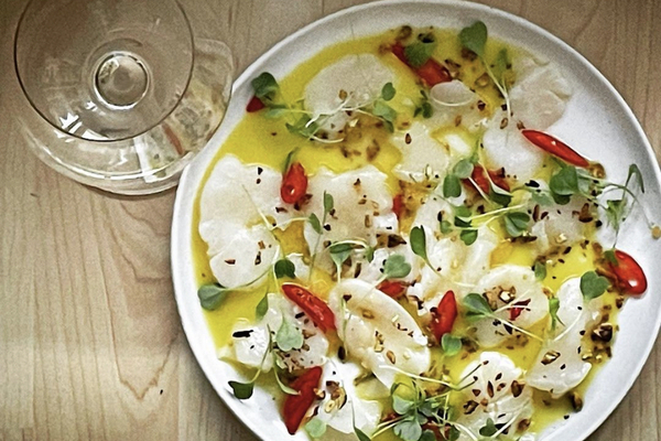 Scallop on a white plate decorated by red chili peppers, micro greens, a toasted pistachios in a pool of lemon oil. Beside the dish is a glass of white wine.