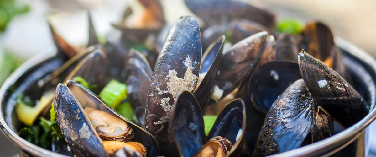 A close up of a large pot of steaming, black mussels flecked with herbs and shallots