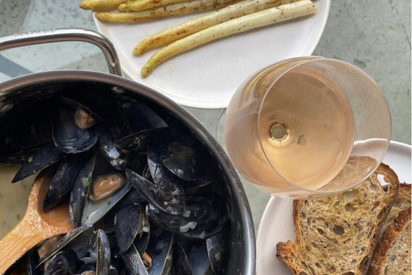 A large pot of black mussels beside a plate of white asparagus, toasted brown bread and a glass of rosÃ©