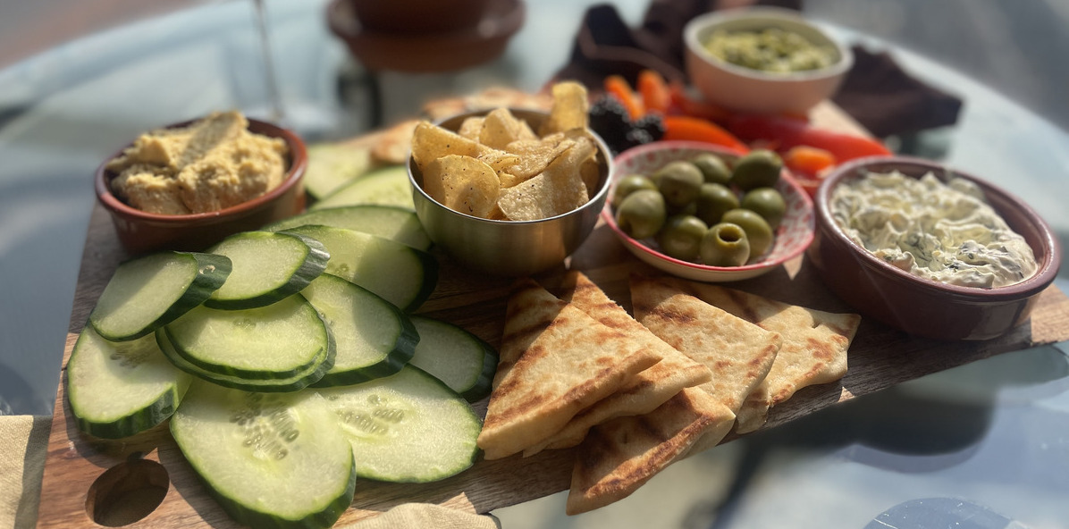 a wood board overflowing with bread, vegetables, olives and three different dips, on a patio table