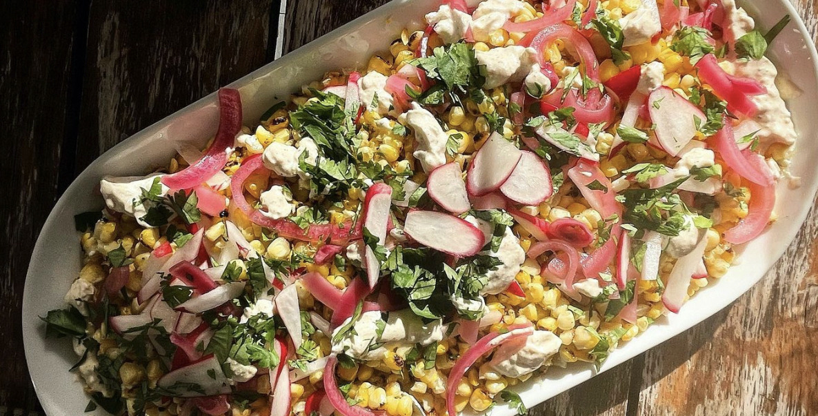 A white platter of corn salad, topped with pink onions, red radishes and green specks of cilantro, sitting on a wood table in the sun