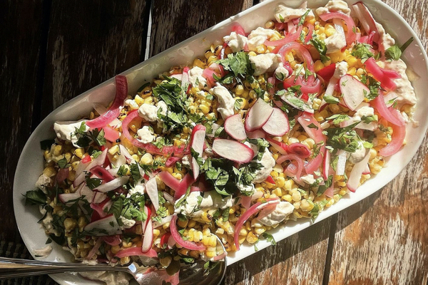 A white platter of corn salad, topped with pink onions, red radishes and green specks of cilantro, sitting on a wood table in the sun