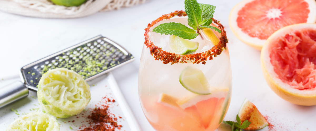 A clear glass of pink paloma with a rusty red salt rim. Beside it is fresh cut red grapefruit and squeezed lime