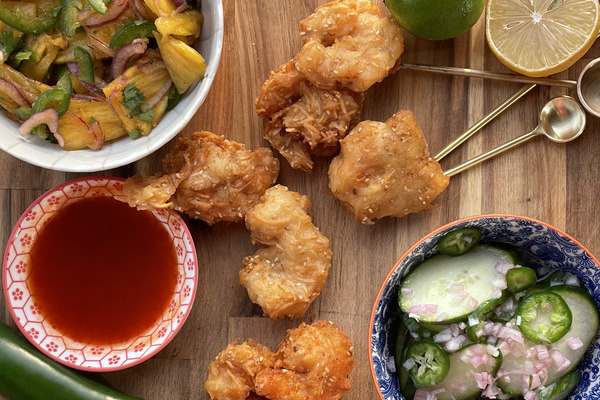 a small pile of coconut shrimp on a wood board surrounded by bowls of cucumber salad, pineapple salad and hot sauce