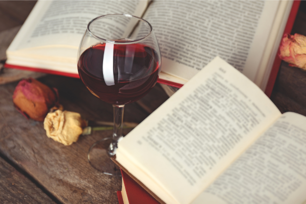 A glass of red wine between two open books on a wood table. Dried roses lay off to the side