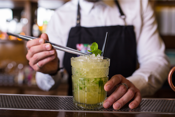 A bartender in an apron placing mint with tweezers in a rocks glass of whisky smash