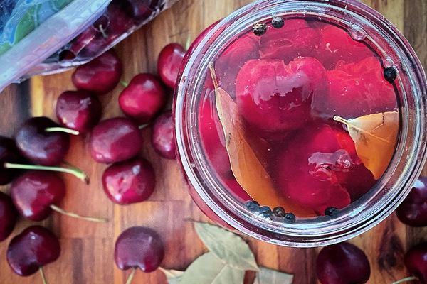 the top view of a jar of pickled cherries with spices, fresh cherries and bay leaves scattered around the jar on a wood board