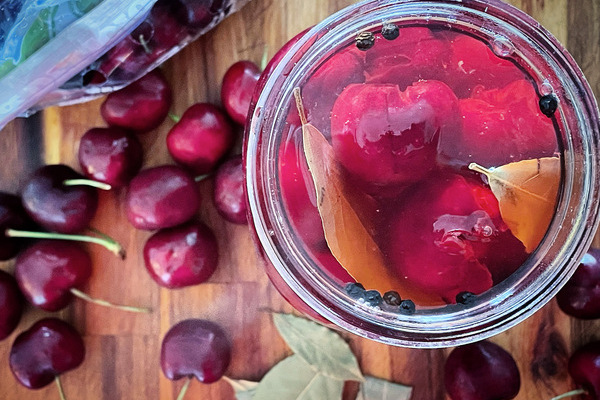 the top view of a jar of pickled cherries with spices, fresh cherries and bay leaves scattered around the jar on a wood board