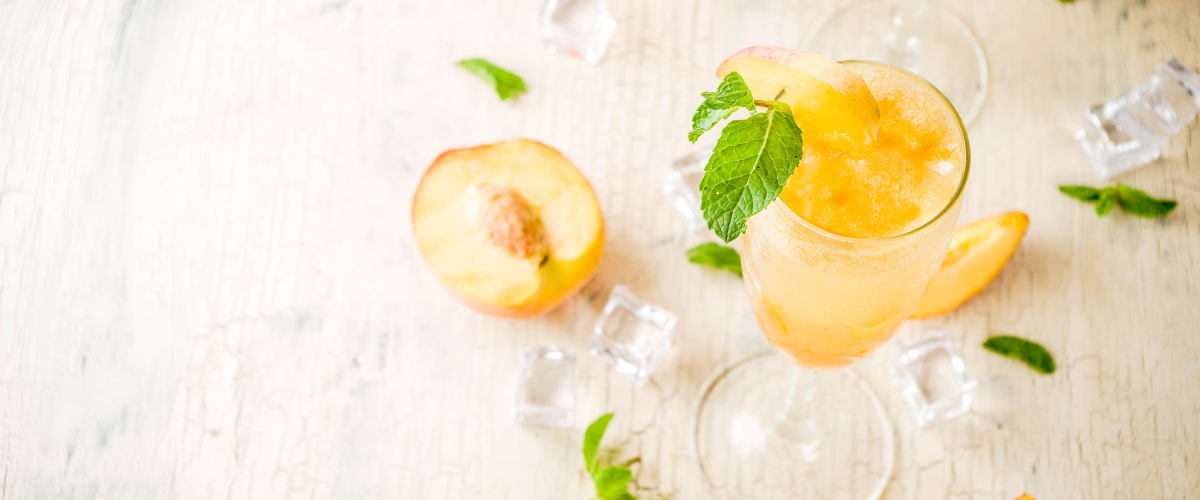 A top view of a flute of Bellini on a creamy white surface with a cut peach beside it and scattering of mint around the glass