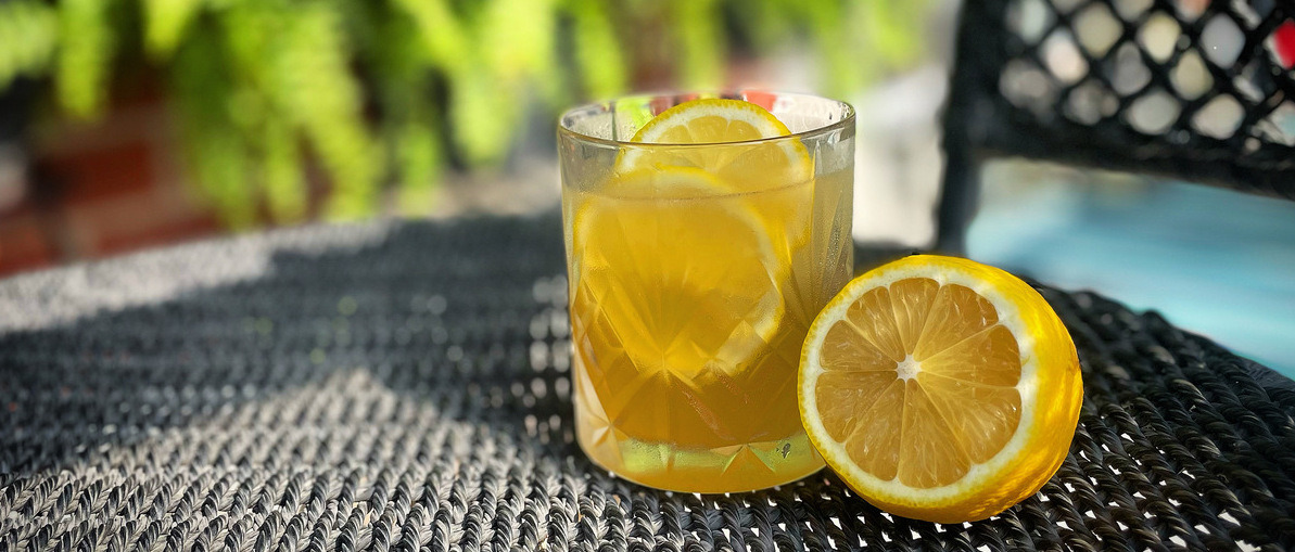 A gold rush cocktail in a rocks glass with lemon slices on a black surface, blurred green plant in the background