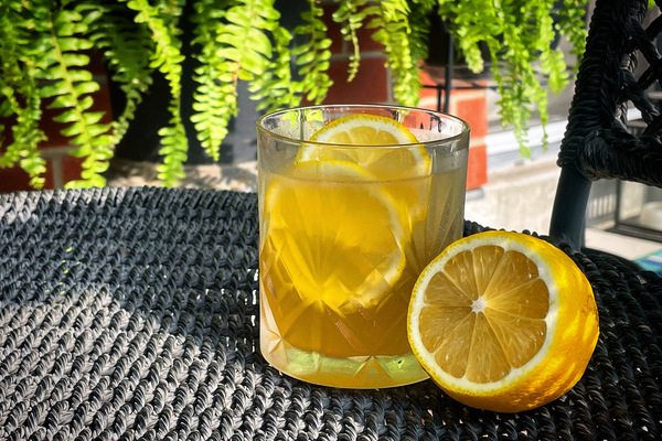 A gold rush cocktail in a rocks glass with lemon slices on a black surface, blurred green plant in the background