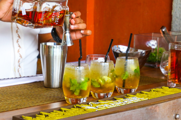 a line of unidentified cocktails on a bar; a pair of hand's pouring unidentified spirit into a shaker