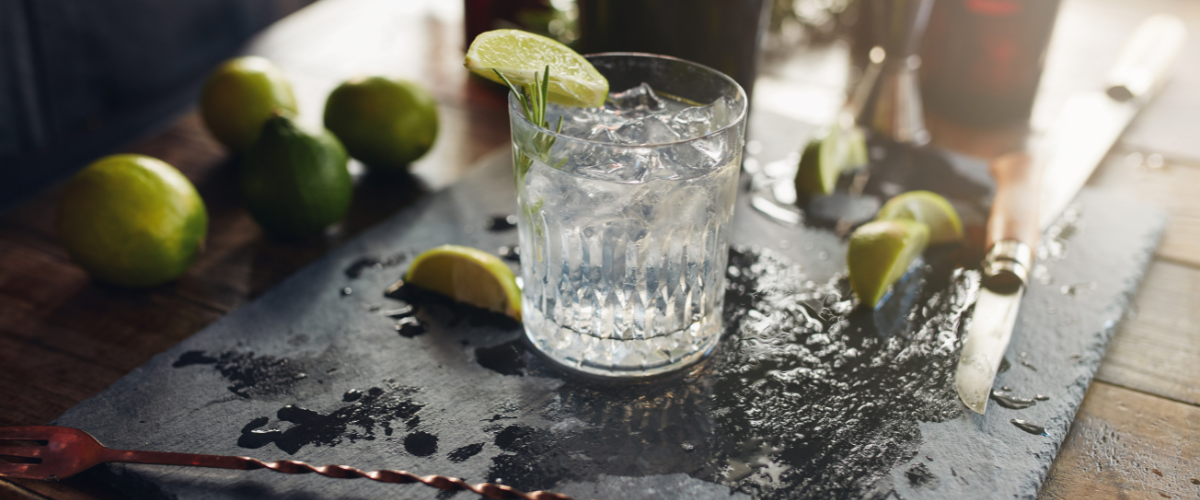 a rocks glass of gin and tonic sitting on a wet black slate board, limes scattered around it