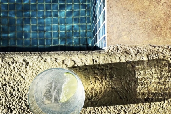A gin and tonic in a rocks glass sitting beside a turquoise blue pool, casting a long shadow.