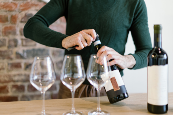A headless man in a green sweater, opening a bottle of wine, 3 empty glasses in front of him