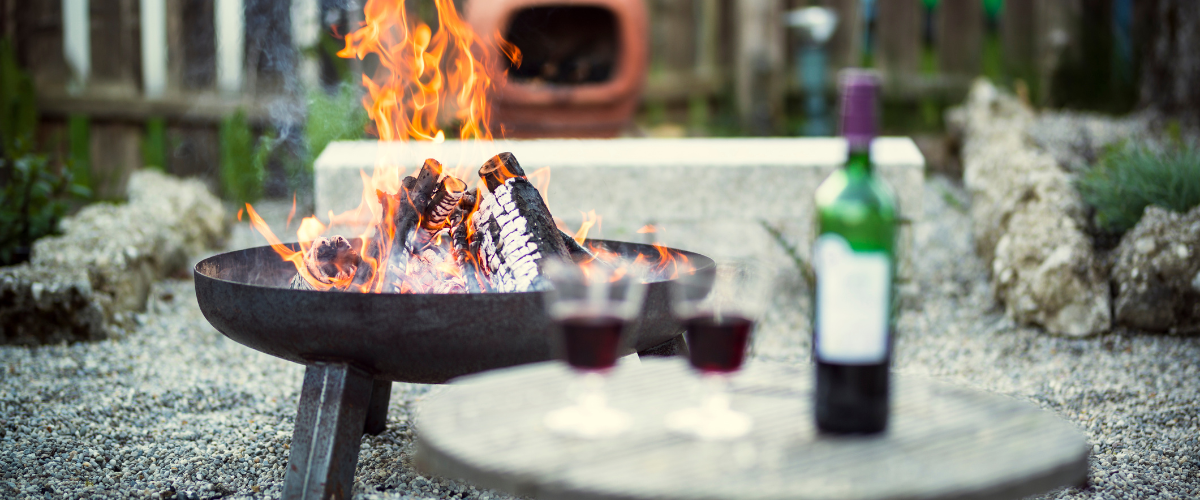 A backyard with a blurry red wine bottle and two rustic wine glasses on a stone table in the foreground, a fire pit in focus, and a clay fire pot in the background