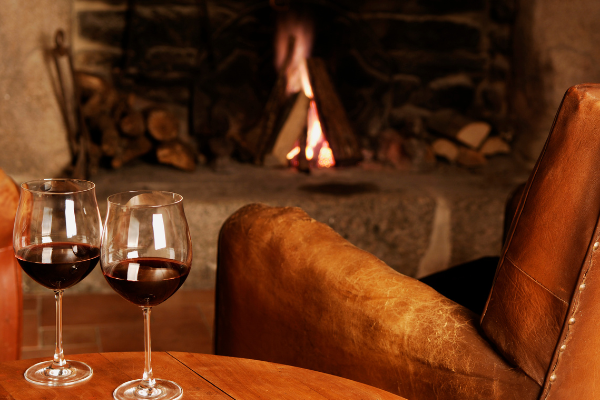 a roaring fire and an empty leather, wing-back chair. Two glasses of red wine on a wood side table.