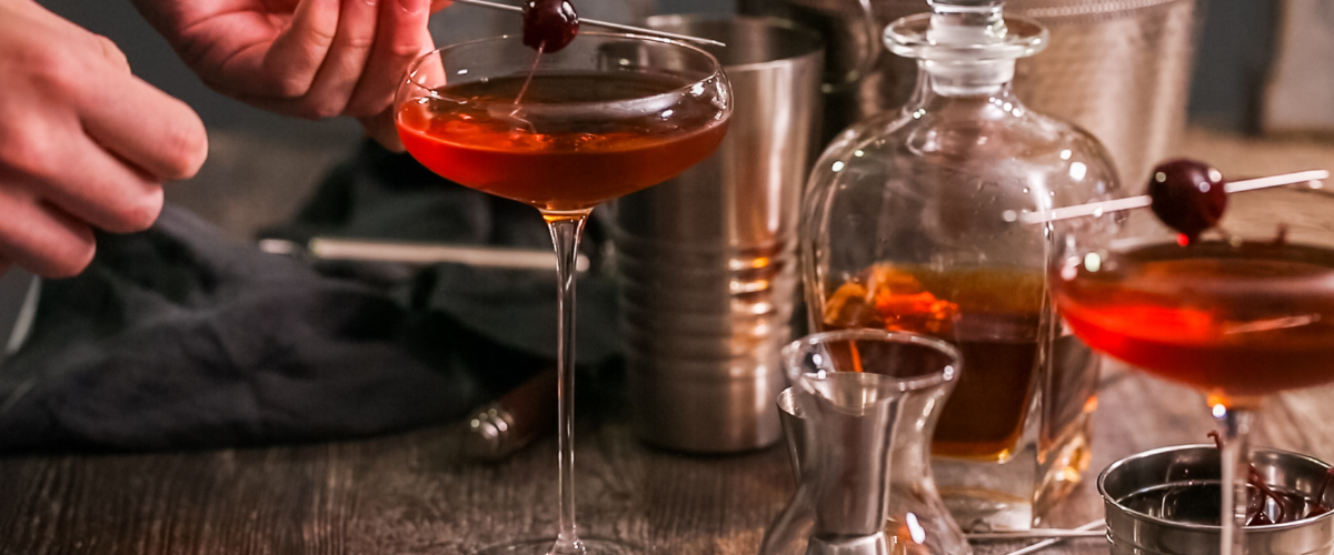 A manhattan cocktail on on a bar, a man's hands placing a speared cherry in the glass