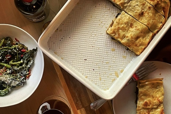 An overview shot of a half empty pan of lasagna, a slice sitting on a white plate beside it, with a bowl of greens off to the side, along with a bottle of red wine a glass of red wine.