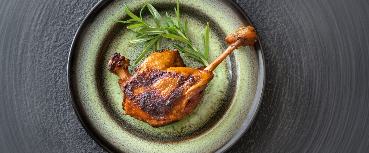 A single duck leg on a beige, clay, round plate with a green tarragon sprig, on a grey, slate table.