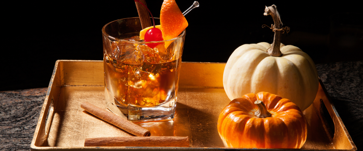 an old fashioned cocktail on a wood tray with an orange and a white pumpkin beside it, along wth cinnamon sticks scattered around the drink