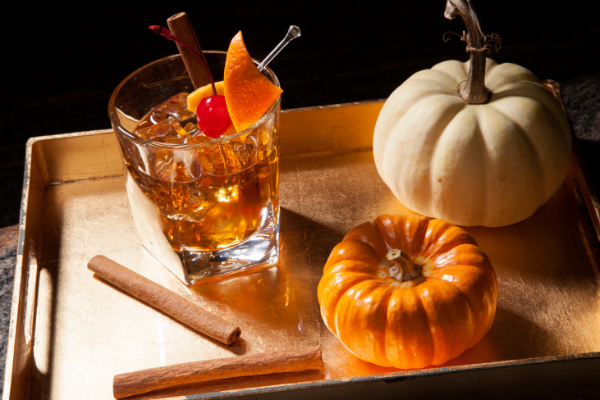 an old fashioned cocktail with an orange and a white pumpkin beside it, along wth cinnamon sticks scattered around the drink