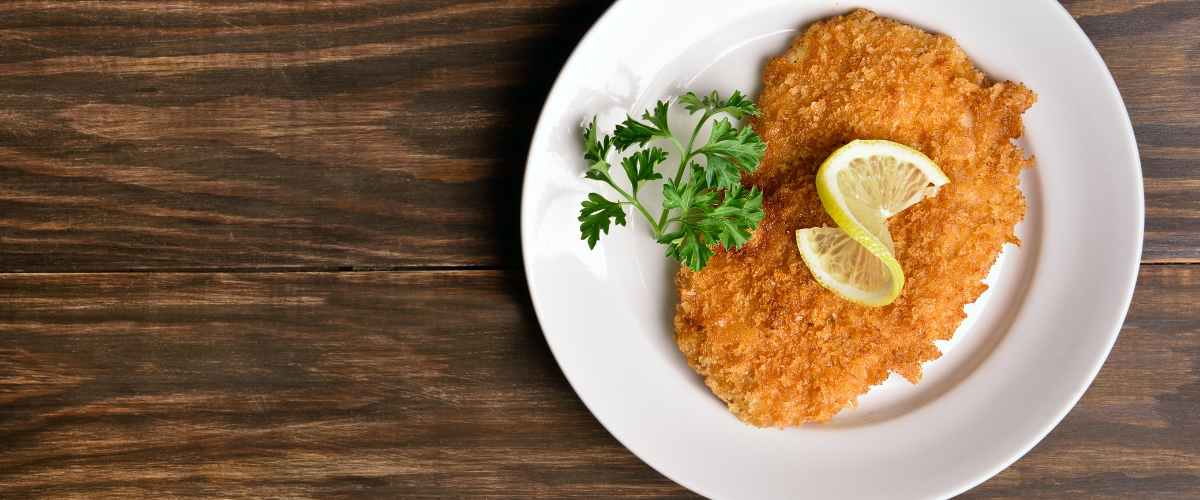 Overhead view of a breaded schnitzel cutlet on a white plate with lemon and parsley