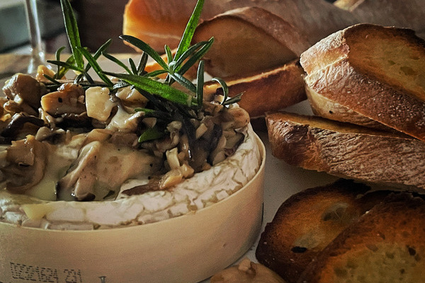 a wheel of camembert in a wood box, topped wth a pile of sauteed mushrooms and rosemary, toasted baguette piled beside it.