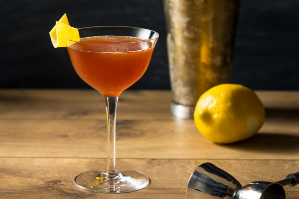 A bright red Paper Plane cocktail in coup with a lemon twist garnish, on a dark wood bar with a lemon and jigger beside it.