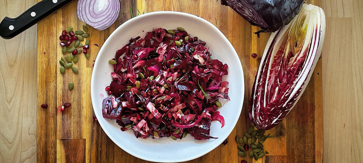A white bowl filled with red and purple mixed vegetables on a wood board. Beside it is a cut purple cabbage, half a red radicchio and half a red onion, pumpkin seeds and pomegranate arils scattered about