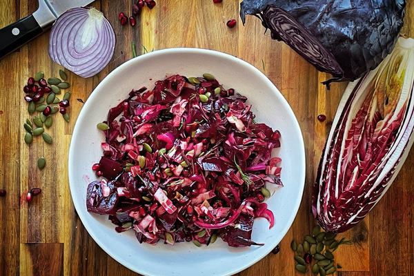 A white bowl filled with red and purple mixed vegetables on a wood board. Beside it is a cut purple cabbage, half a red radicchio and half a red onion, pumpkin seeds and pomegranate arils scattered about.
