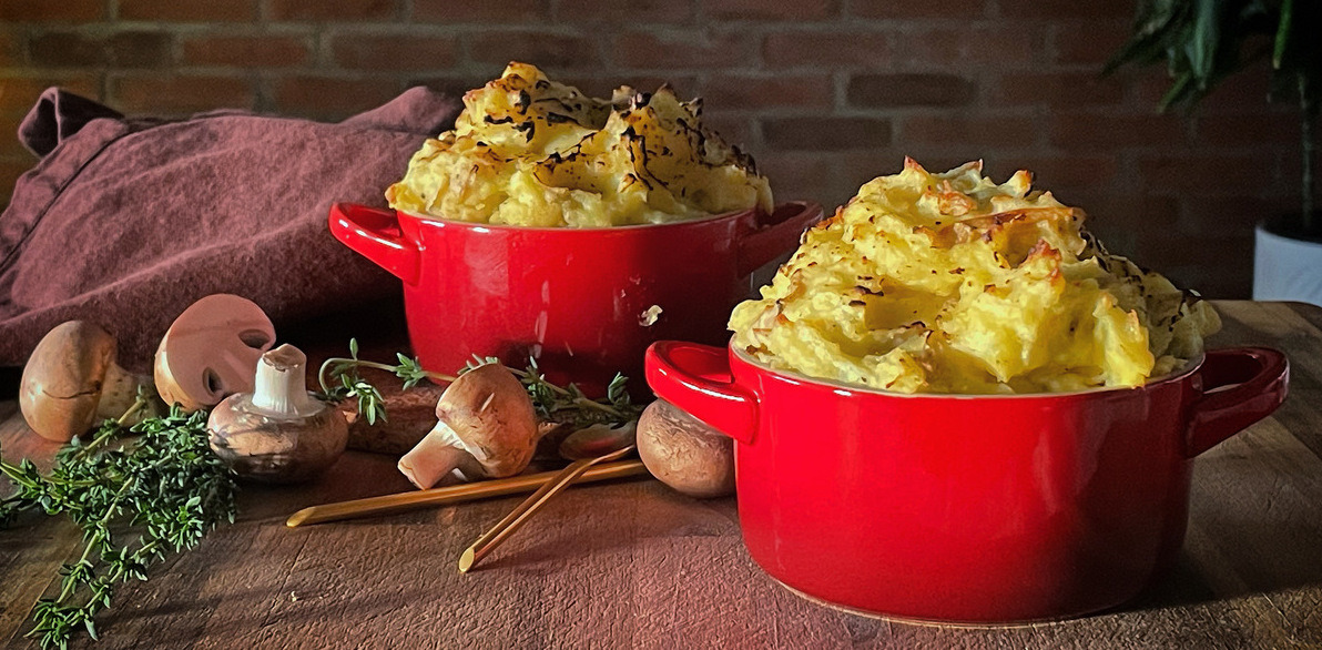 Two red ramekins topped with crusty mashed potato on a wooden board with scattered thyme and mushrooms