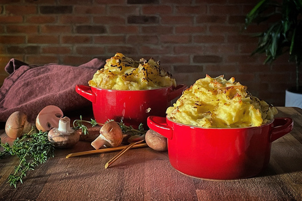 Two red ramekins topped with crusty mashed potato on a wooden board with scattered thyme and mushrooms