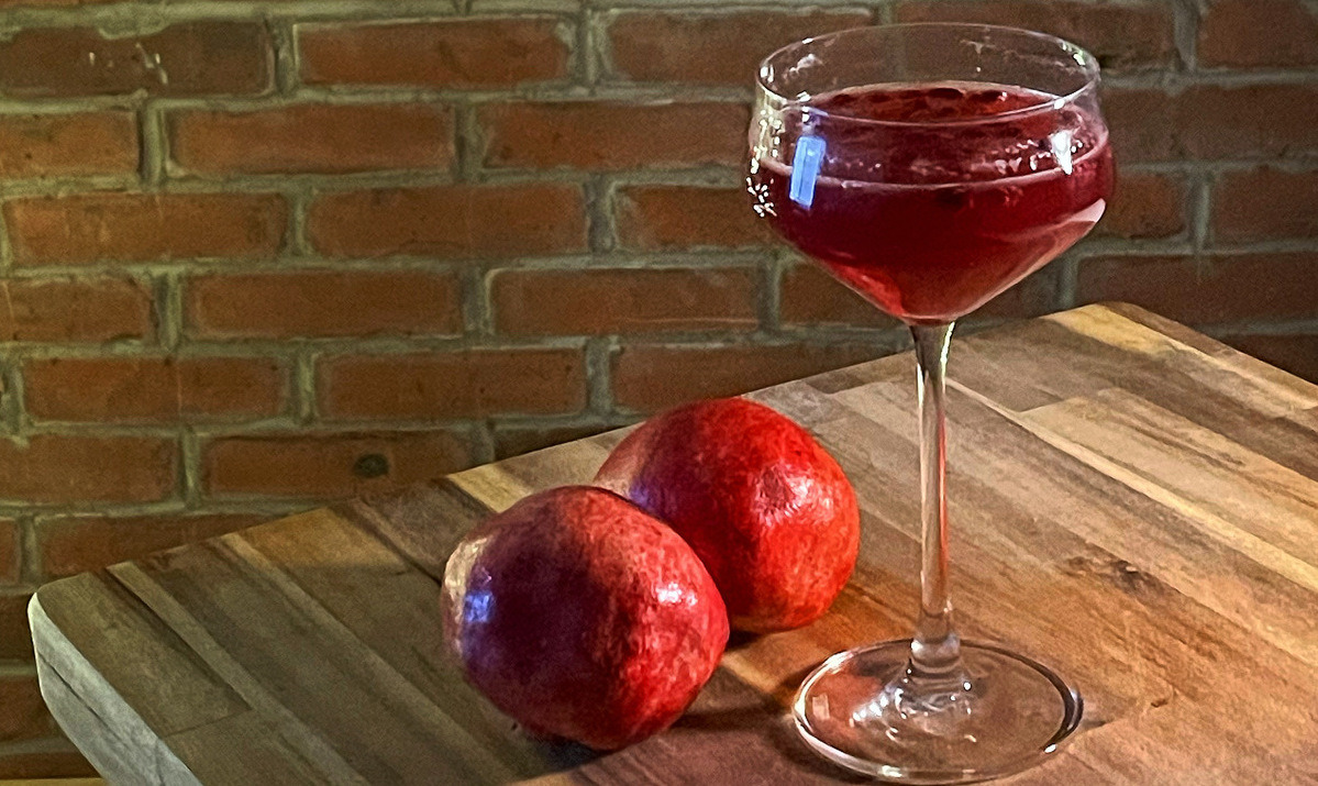 A coup fill with red liquid on a wood board, two pomegranates beside it. A brick wall in the background.